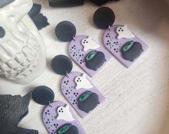 May include: Halloween-themed earrings featuring a black circle stud, a lavender arch with a white ghost, a black cauldron, and green dots. The earrings are displayed on a white wooden surface.