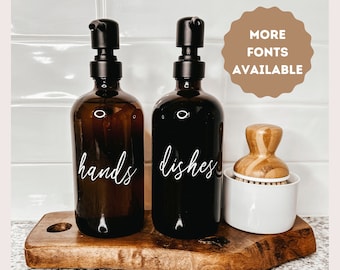 May include: Two amber glass pump bottles with black pumps and white labels that say "hands" and "dishes". The bottles are on a wooden tray with a white ceramic dish and a wooden brush.