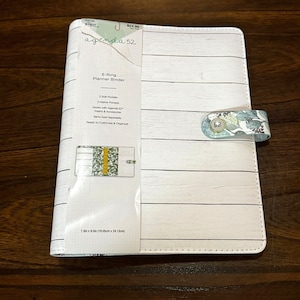 May include: White 6-ring planner binder with a floral design and a white woodgrain pattern. The binder has two side pockets and three interior pockets. It is 7.5 inches by 9.5 inches (19.05 cm x 24.13 cm).