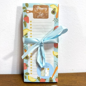 May include: A blue and white notepad with a shopping list design. The notepad has a light blue ribbon tied around it. The notepad has a white background with a yellow and orange fruit and vegetable design. The notepad has the words "Shopping List" written on it in black.