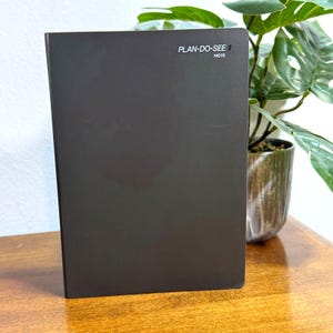 May include: A dark gray notebook with the text "PLAN-DO-SEE NOTE" in white. The notebook is standing upright on a wooden surface, with a potted plant in the background.