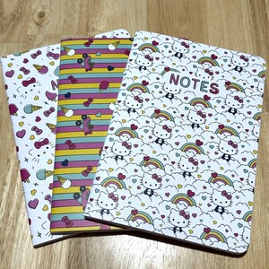 May include: Three notebooks with colorful patterned covers. The front cover of the notebook on the right has a white background with a repeating pattern of pink hearts, rainbows, and cartoon Hello Kitty characters. The text "NOTES" is printed in black on the front cover. The notebook in the middle has a pink, blue, and yellow striped cover with a repeating pattern of hearts, ice cream cones, and roller skates. The notebook on the left has a white background with a repeating pattern of pink hearts, blue stars, and cartoon Hello Kitty characters.