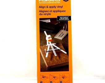 May include: White vinyl alignment tool with orange accents. The tool is shown on a book with the words "adventure" and "born to travel" on it. The packaging is orange and white with the Fiskars logo and the words "Align & apply vinyl".