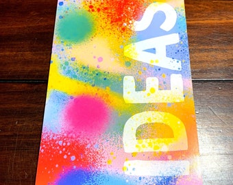 Puede incluir: Un cuaderno colorido con un diseño de pintura en aerosol y la palabra "IDEAS" en letras blancas.