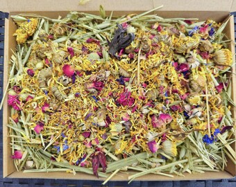 May include: A box filled with dried flowers and herbs, including yellow, pink, and blue petals, and green stems. The box is made of cardboard and has a brown color.