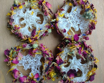 May include: Four circular, white, textured objects, each adorned with dried flower petals in shades of pink, yellow, and blue. The objects have a unique, open-weave design, and are arranged on a light brown wooden surface.
