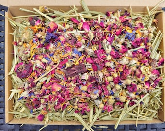May include: A box filled with a mixture of dried flowers and herbs, including pink, red, yellow, and blue petals. The mixture is in a brown cardboard box with a light brown straw-like material.