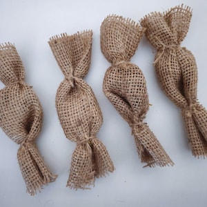 Op de afbeelding: Vier bruine jute stoffen cadeauzakken die bovenaan met een knoop zijn vastgebonden. De zakken zijn in een rij op een wit oppervlak gerangschikt.