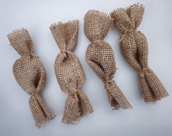 May include: Four brown burlap fabric gift bags tied with a knot at the top. The bags are arranged in a row on a white surface.
