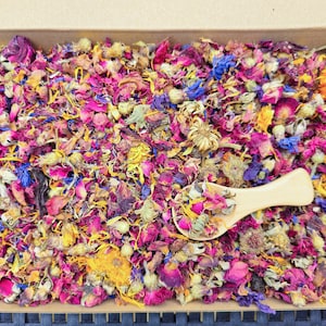 Pode incluir: Uma caixa cheia de uma mistura de pétalas de flores secas em tons de rosa, roxo, amarelo e laranja. Uma pequena colher de madeira está descansando sobre as pétalas.