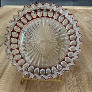 Puede incluir: Un plato para pastel de vidrio transparente con un borde festoneado y un borde rojo. El plato tiene un patr&oacute;n decorativo de l&iacute;neas radiantes y peque&ntilde;os puntos en relieve alrededor del borde.