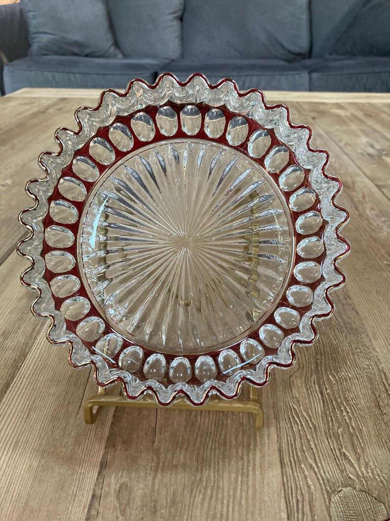Puede incluir: Un plato para pastel de vidrio transparente con un borde festoneado y un borde rojo. El plato tiene un patr&oacute;n decorativo de l&iacute;neas radiantes y peque&ntilde;os puntos en relieve alrededor del borde.