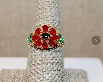 Puede incluir: Un anillo de tono dorado con una piedra ovalada negra central rodeada por un halo de piedras rojas en forma de pera dispuestas como pétalos de flor. Los lados están acentuados con pequeñas piedras verdes, creando un diseño floral multicolor.