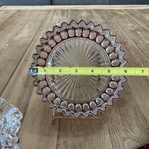 Puede incluir: Un plato de vidrio transparente con un borde festoneado y un borde rojo. El plato tiene un patr&oacute;n decorativo de peque&ntilde;os c&iacute;rculos alrededor del borde y un patr&oacute;n radial en el centro. El plato est&aacute; sobre una superficie de madera y se est&aacute; midiendo con una cinta m&eacute;trica.