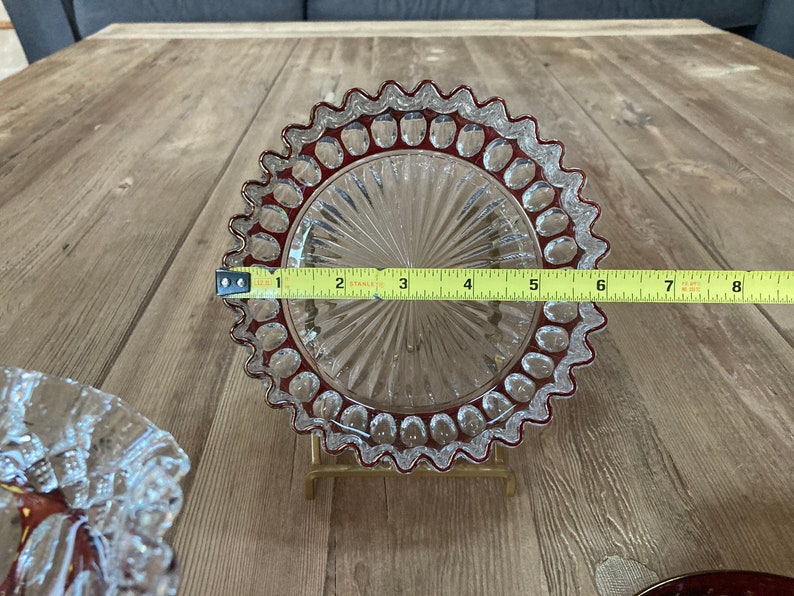 Puede incluir: Un plato de vidrio transparente con un borde festoneado y un borde rojo. El plato tiene un patr&oacute;n decorativo de peque&ntilde;os c&iacute;rculos alrededor del borde y un patr&oacute;n radial en el centro. El plato est&aacute; sobre una superficie de madera y se est&aacute; midiendo con una cinta m&eacute;trica.
