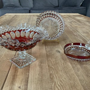 Puede incluir: Tres cuencos de vidrio transparente con un tinte rojo rub&iacute;. Los cuencos tienen un borde festoneado y un patr&oacute;n decorativo. Los cuencos est&aacute;n sobre una superficie de madera.