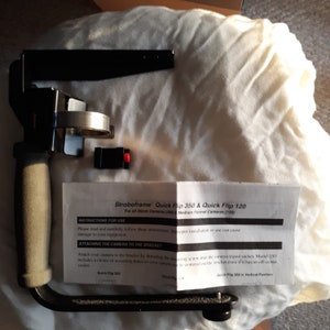 May include: Black camera bracket with a handle and a silver knob. The bracket is designed to hold a camera in a vertical position. The instructions for use are printed on a white paper with the text "Stroboframe Quick Flip 350 & Quick Flip 120".