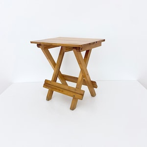 May include: A wooden folding stool with a natural finish. The stool is made of solid wood and has a simple, classic design.