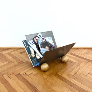 Peut inclure: Un porte-revues en métal moderne avec un design géométrique, soutenu par deux sphères en bois. Le porte-revues contient plusieurs magazines de mode, dont Vogue et Elle, sur un sol en bois.