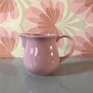 Puede incluir: Una jarra de cerámica de color rosa pálido con cuerpo redondeado y asa. La jarra está sobre una superficie gris.