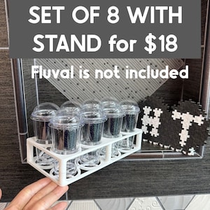 May include: White plastic stand for eight small clear containers with black filter media inside. The stand is mounted to a wall. The text "SET OF 8 WITH STAND for $18 Fluval is not included" is displayed above the stand.