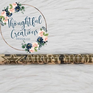 Pode incluir: Caneta com padr&atilde;o de camuflagem e as palavras "ARMY MOM" em letras douradas. A caneta tem um design de estrela e uma ponta preta. O fundo &eacute; uma superf&iacute;cie macia e branca.
