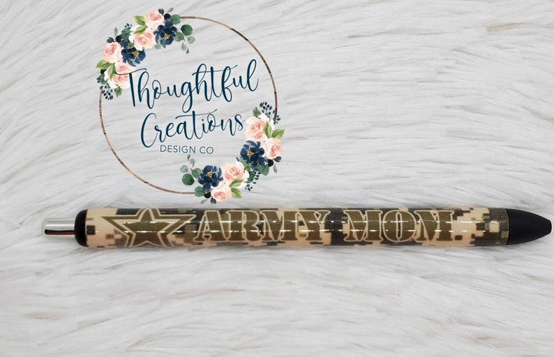Pode incluir: Caneta com padr&atilde;o de camuflagem e as palavras "ARMY MOM" em letras douradas. A caneta tem um design de estrela e uma ponta preta. O fundo &eacute; uma superf&iacute;cie macia e branca.