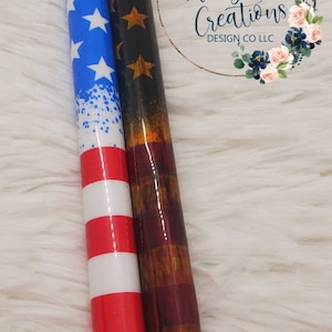 Pode incluir: Duas canetas com designs patri&oacute;ticos e detalhes prateados. Uma apresenta um design de estrelas azuis e brancas, a outra um design vermelho, branco e castanho. As canetas t&ecirc;m um acabamento brilhante e pontas azuis e pretas. Medem cerca de 15 cm.