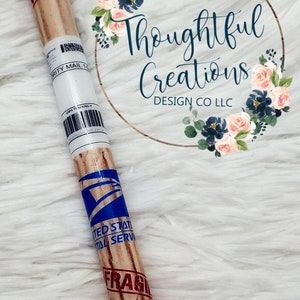 Pode incluir: Caneta azul e castanha com tema USPS. A caneta tem uma ponta azul e um corpo projetado para se parecer com um pacote, com a palavra "FRAGILE" e "UNITED STATES POSTAL SERVICE" impressas. A caneta &eacute; decorada com um design floral.