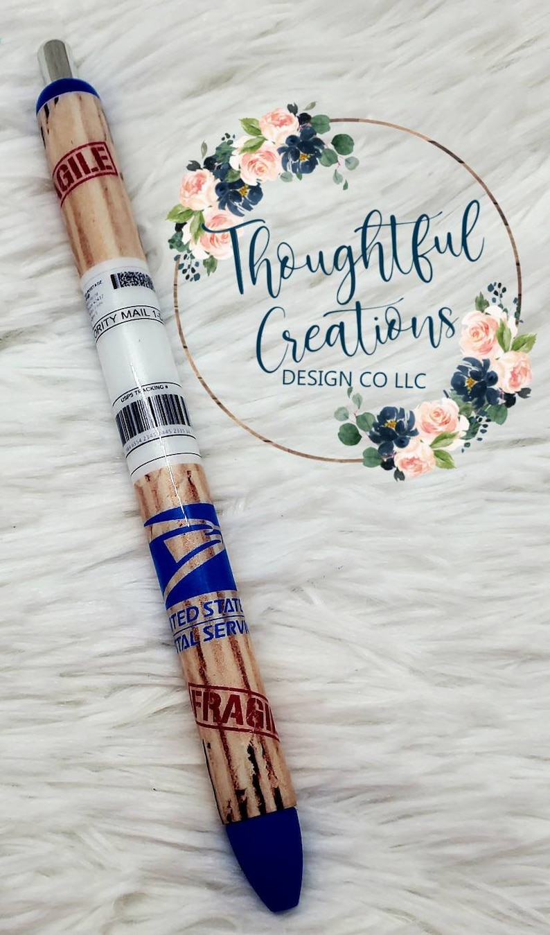 Pode incluir: Caneta azul e castanha com tema USPS. A caneta tem uma ponta azul e um corpo projetado para se parecer com um pacote, com a palavra "FRAGILE" e "UNITED STATES POSTAL SERVICE" impressas. A caneta &eacute; decorada com um design floral.
