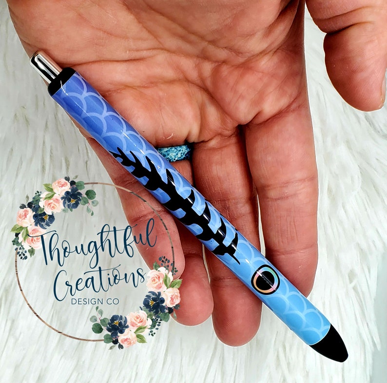 Pode incluir: Caneta azul com padr&atilde;o de escamas e detalhes em preto. A caneta apresenta um design preto e o texto "Thoughtful Creations Design Co.". Possui uma ponta prateada e uma ponta preta.