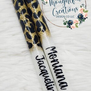 Pode incluir: Duas canetas personalizadas com um design de estampa de leopardo preto e dourado. As canetas t&ecirc;m uma base branca com glitter com os nomes "Jacqueline" e "Montana" em escrita preta. As canetas t&ecirc;m pontas pretas e detalhes prateados.