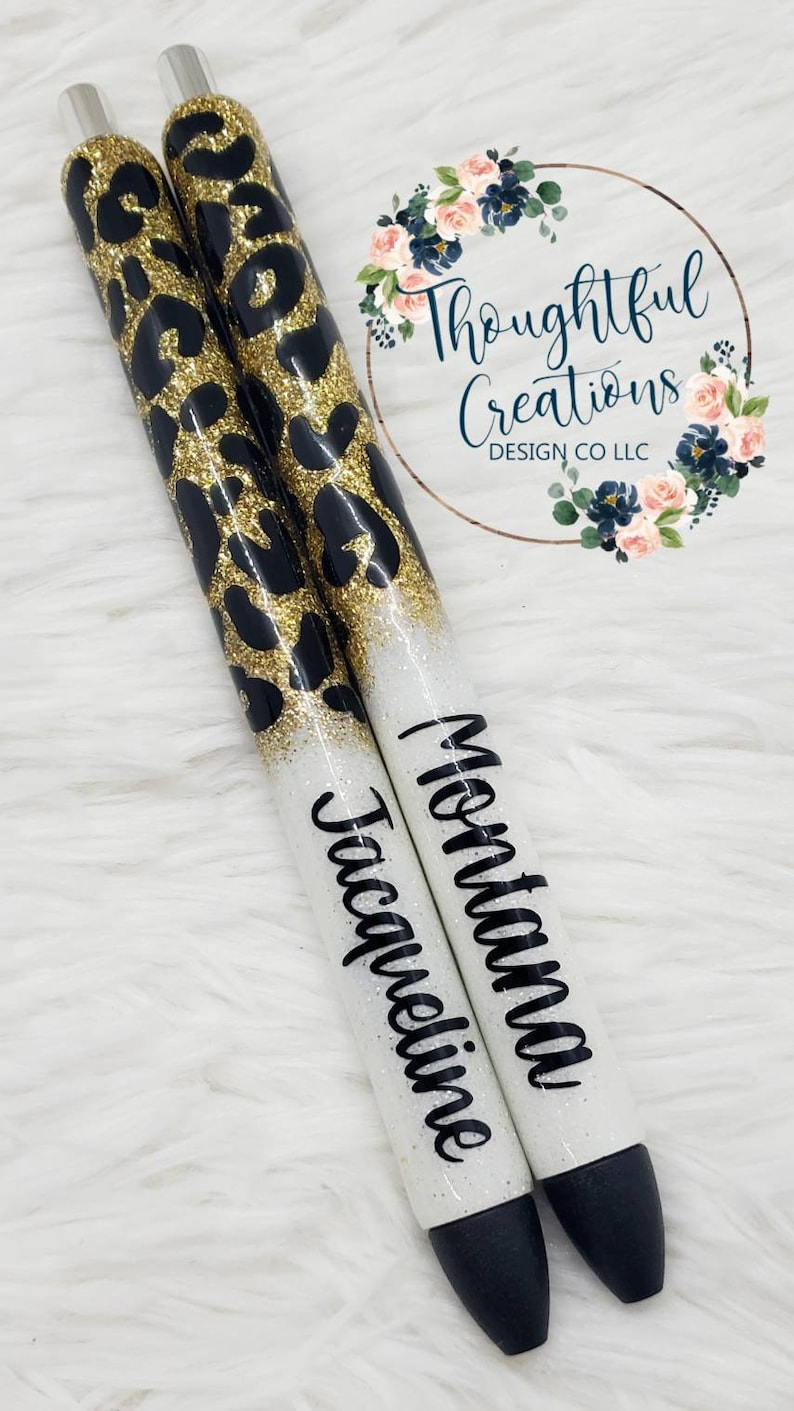 Pode incluir: Duas canetas personalizadas com um design de estampa de leopardo preto e dourado. As canetas t&ecirc;m uma base branca com glitter com os nomes "Jacqueline" e "Montana" em escrita preta. As canetas t&ecirc;m pontas pretas e detalhes prateados.