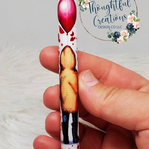 Pode incluir: Caneta branca com um bal&atilde;o vermelho e uma figura com capa de chuva amarela. A caneta tem respingos vermelhos e uma ponta vermelha. A caneta &eacute; segurada em uma m&atilde;o.