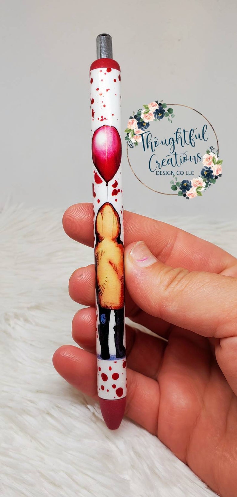 Pode incluir: Caneta branca com um bal&atilde;o vermelho e uma figura com capa de chuva amarela. A caneta tem respingos vermelhos e uma ponta vermelha. A caneta &eacute; segurada em uma m&atilde;o.