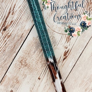 May include: A glittery teal pen with a silver tip and a black ink cartridge. The pen's barrel transitions to a cowhide pattern. The pen is set against a light wood grain background. The text "Thoughtful Creations Design Co LLC" is visible.