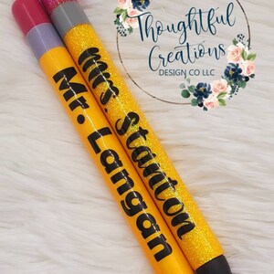 Pode incluir: Duas canetas personalizadas com detalhes de glitter. Uma caneta &eacute; amarela com "Mr. Langan" em preto, a outra &eacute; amarela com "Mrs. Stanton" em preto. Ambas as canetas t&ecirc;m pontas pretas e tampas prateadas. As canetas est&atilde;o sobre uma superf&iacute;cie branca e fofa.