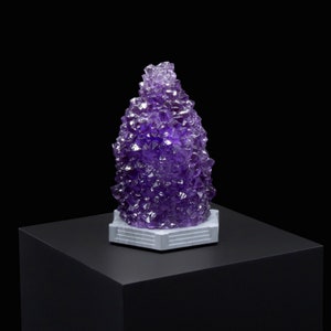 May include: A purple amethyst crystal cluster on a white hexagonal base. The crystal is a tall, pointed shape and is made up of many smaller crystals.