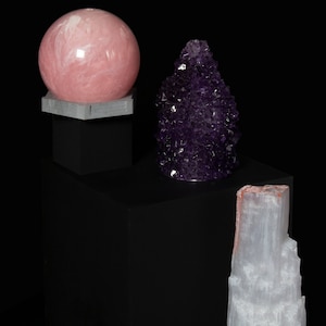 Pode incluir: Três formações de cristal em plataformas pretas. Uma grande esfera rosa, um aglomerado de ametista roxa e uma torre de selenita branca.