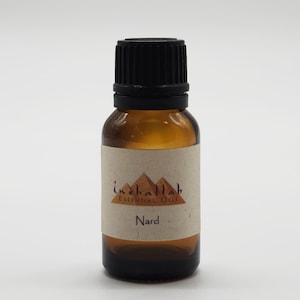 May include: A small amber glass bottle with a black cap, labeled "inaballah Essential Oils" and "Nard." The label features a mountain graphic. The bottle contains essential oil.
