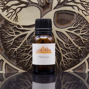 Może przedstawiać: Mała, brązowa szklana butelka olejku eterycznego Heliotrop z czarną zakrętką. Etykieta zawiera tekst "inaballah essential oils" i "Heliotrope". Butelka stoi na drewnianym tle z motywem drzewa.