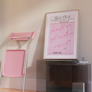 May include: A pink folding chair with a black metal frame sits next to a black side table with a framed pink guest check that reads "should we get another round?"