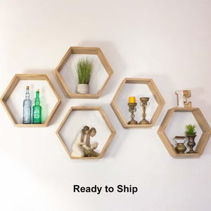 May include: Five wooden hexagon shelves mounted on a white wall, each displaying decorative items. The shelves hold items like glass bottles, candles, figurines, and small plants. The text "Ready to Ship" is at the bottom.