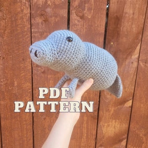 May include: A gray crocheted manatee plush toy with black eyes. The toy is being held in a hand against a wooden background. The text "PDF PATTERN" is written in white letters on a yellow background.