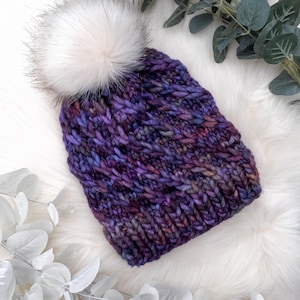 May include: A chunky-knit beanie hat in shades of purple, blue, and pink, with a large, fluffy white faux fur pom-pom. The hat is displayed on a white, textured surface with green and white foliage accents.