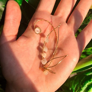 May include: A gold wire ear cuff with a delicate design featuring a pink stone and smaller white stones. The cuff is shown on a hand against a green leafy background.