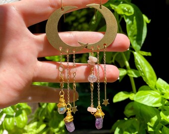 May include: Gold crescent moon earrings with dangling charms. The charms include amethyst crystals, gold stars, and small pink and clear beads.