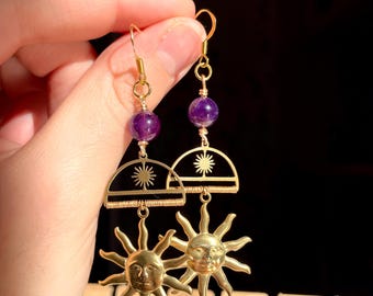 May include: Gold earrings with a sun design and a purple amethyst bead. The sun has a face and rays.
