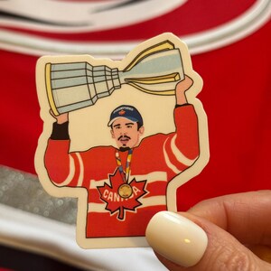 May include: A sticker of a hockey player wearing a red and white jersey with a maple leaf and the word "CANADA" on it. He is holding a trophy above his head with the word "CHAMPIONS" on it. He is wearing a gold medal around his neck.