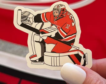Puede incluir: Una pegatina que representa a un portero de hockey con uniforme rojo y negro, con el número 32. El portero está agachado, sosteniendo un palo de hockey y usando un casco. La pegatina está sobre un fondo rojo.